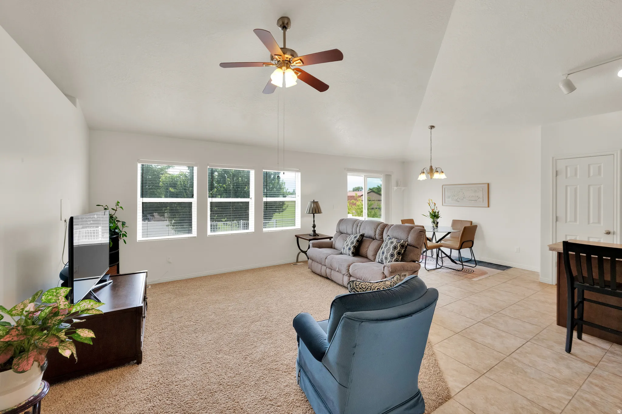 Spacious living area featuring vaulted ceilings with a ceiling fan, three large windows, and carpet flooring