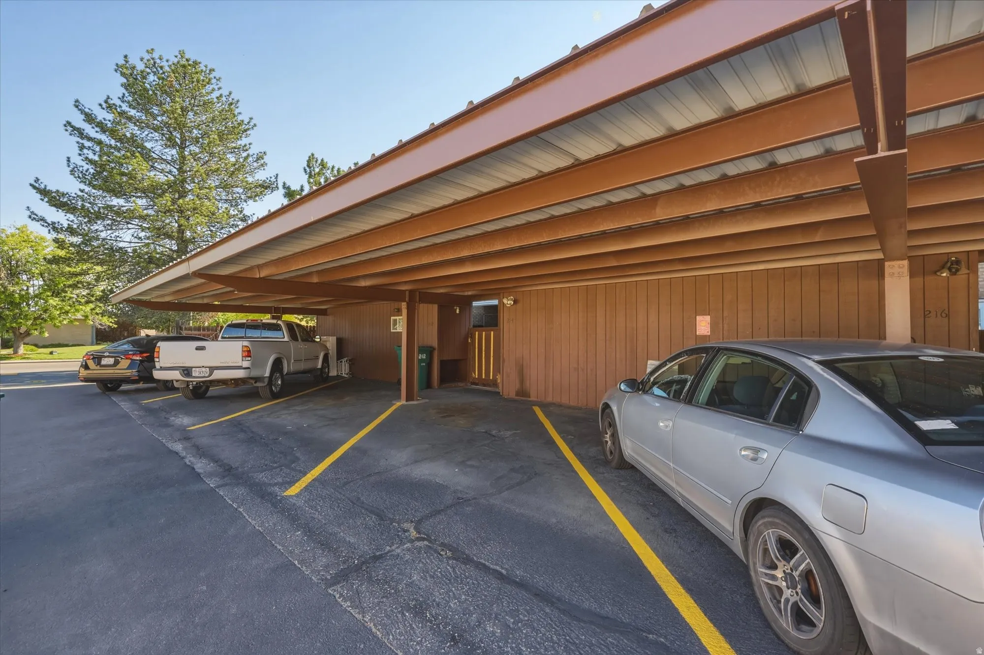 Covered parking structure featuring a metal roof, exposed brown beams, wood-paneled walls, and asphalt pavement with designated parking spaces