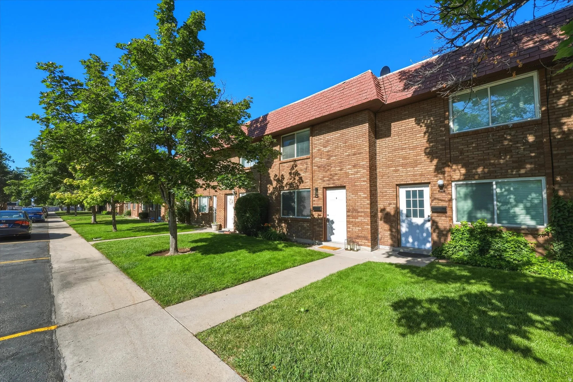 Brick exterior townhouse featuring a red tile roof, white entry doors, and mature trees lining the sidewalk