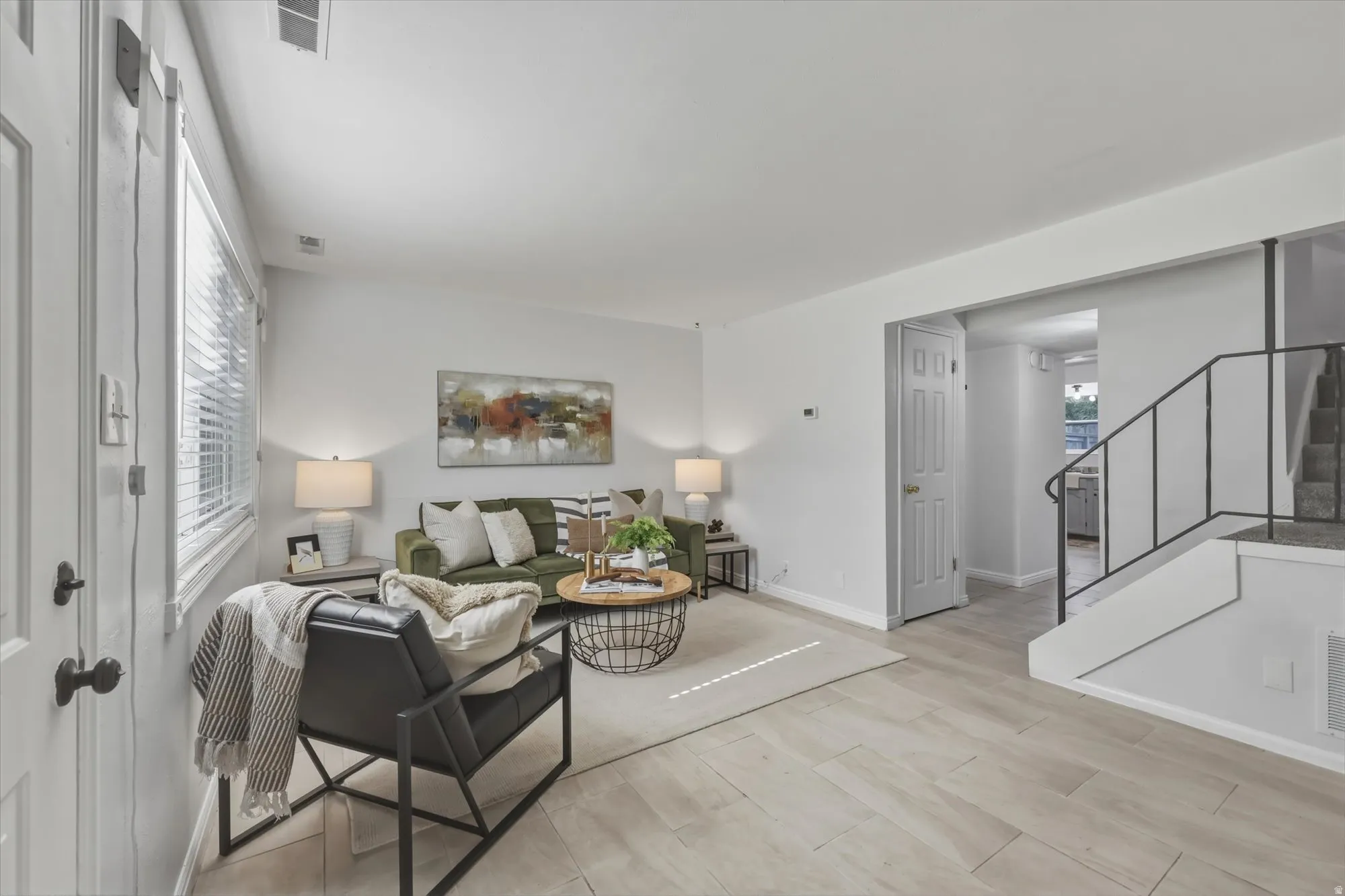 Spacious living area featuring white walls, and a large window with horizontal blinds. Stairs lead to second floor.