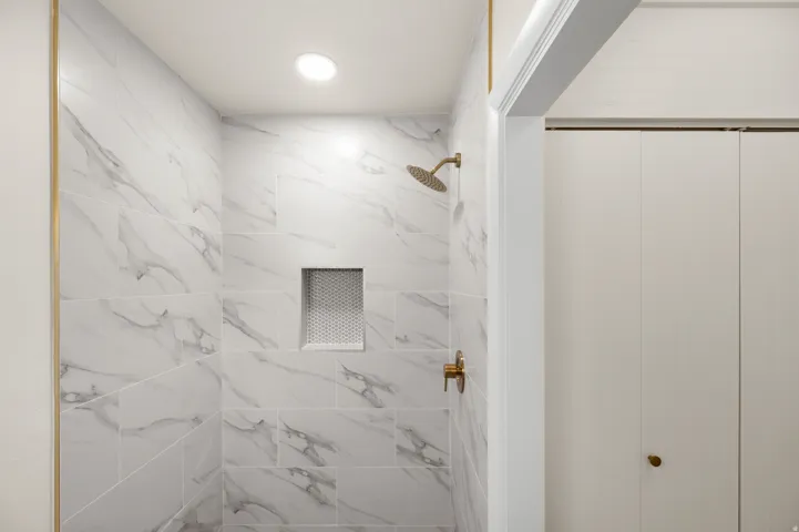 Shower with white marble-patterned tile, gold-finish fixtures, recessed lighting, and a built-in shower niche with a textured back wall