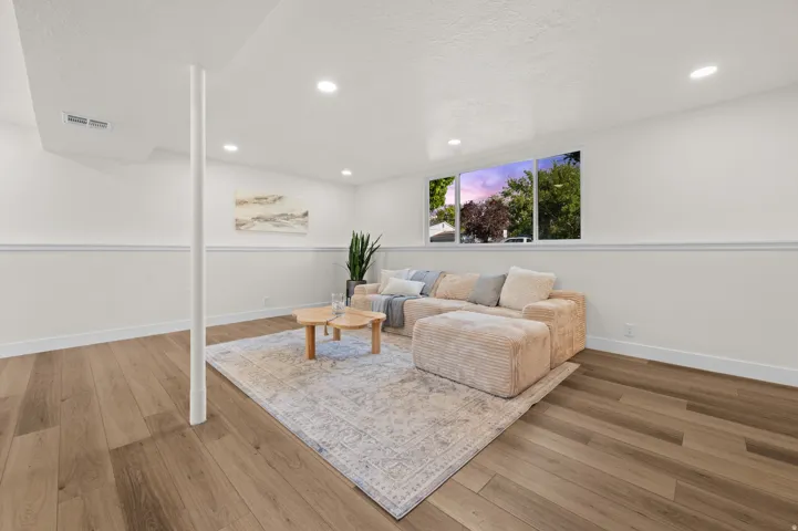 Recessed lighting illuminates this bright living space featuring wood-finish flooring, a white pillar, and a large window