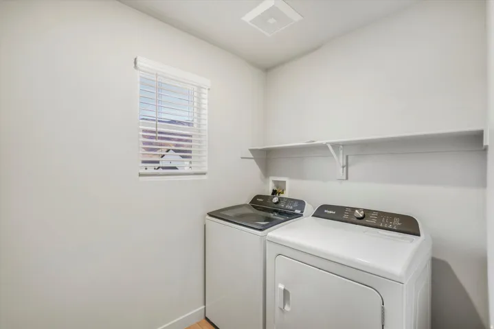 Dedicated laundry area featuring a window with white blinds, a white storage shelf, light gray walls, and wood-finish flooring