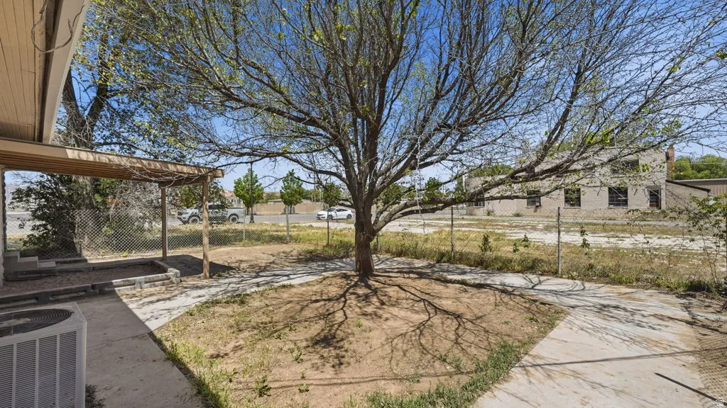 Large mature tree providing shade, concrete pathway, covered patio structure, chain-link fencing, and an exposed aggregate porch