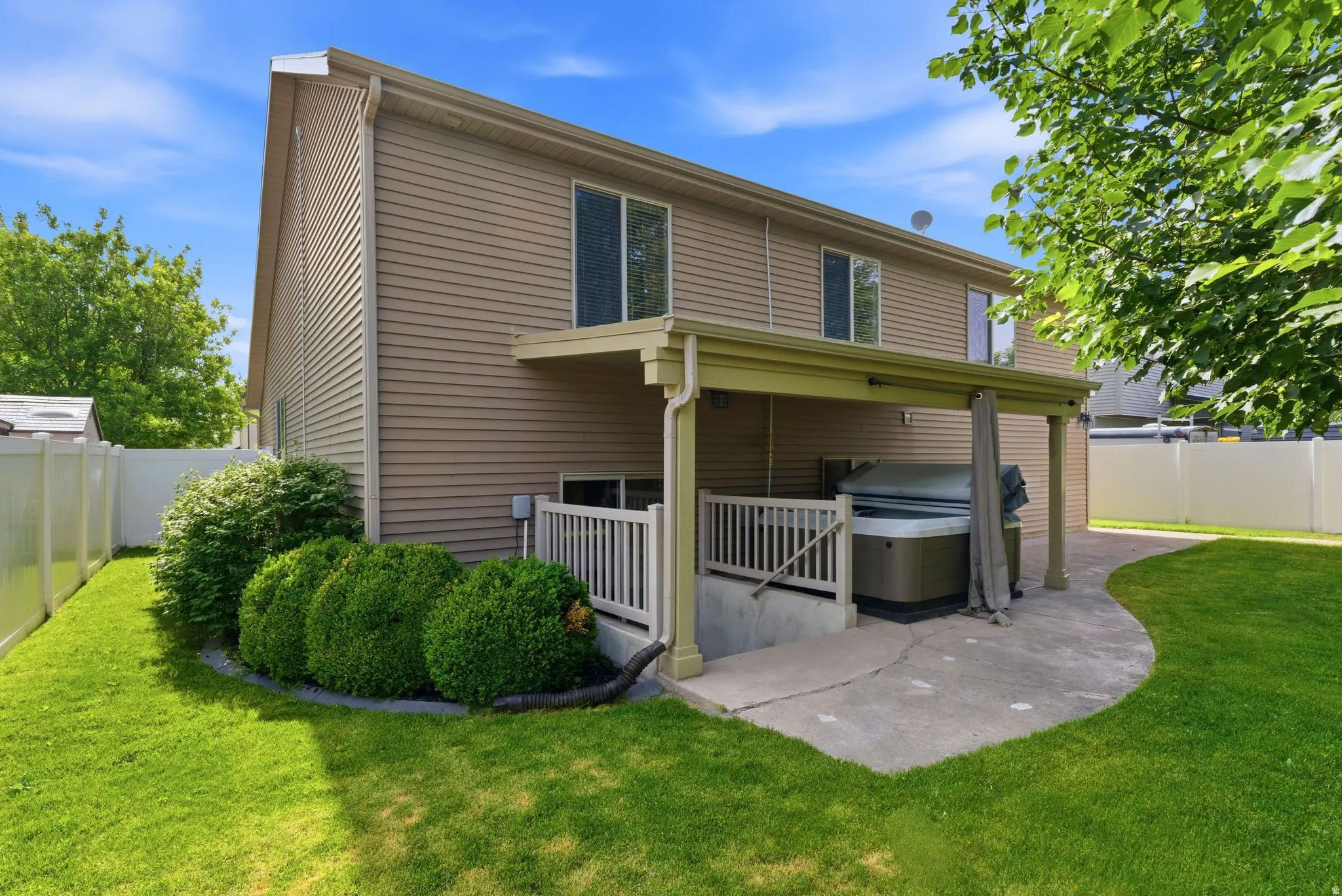 Rear exterior featuring a covered patio,  hot tub, concrete walkway, vibrant lawn, and privacy fencing