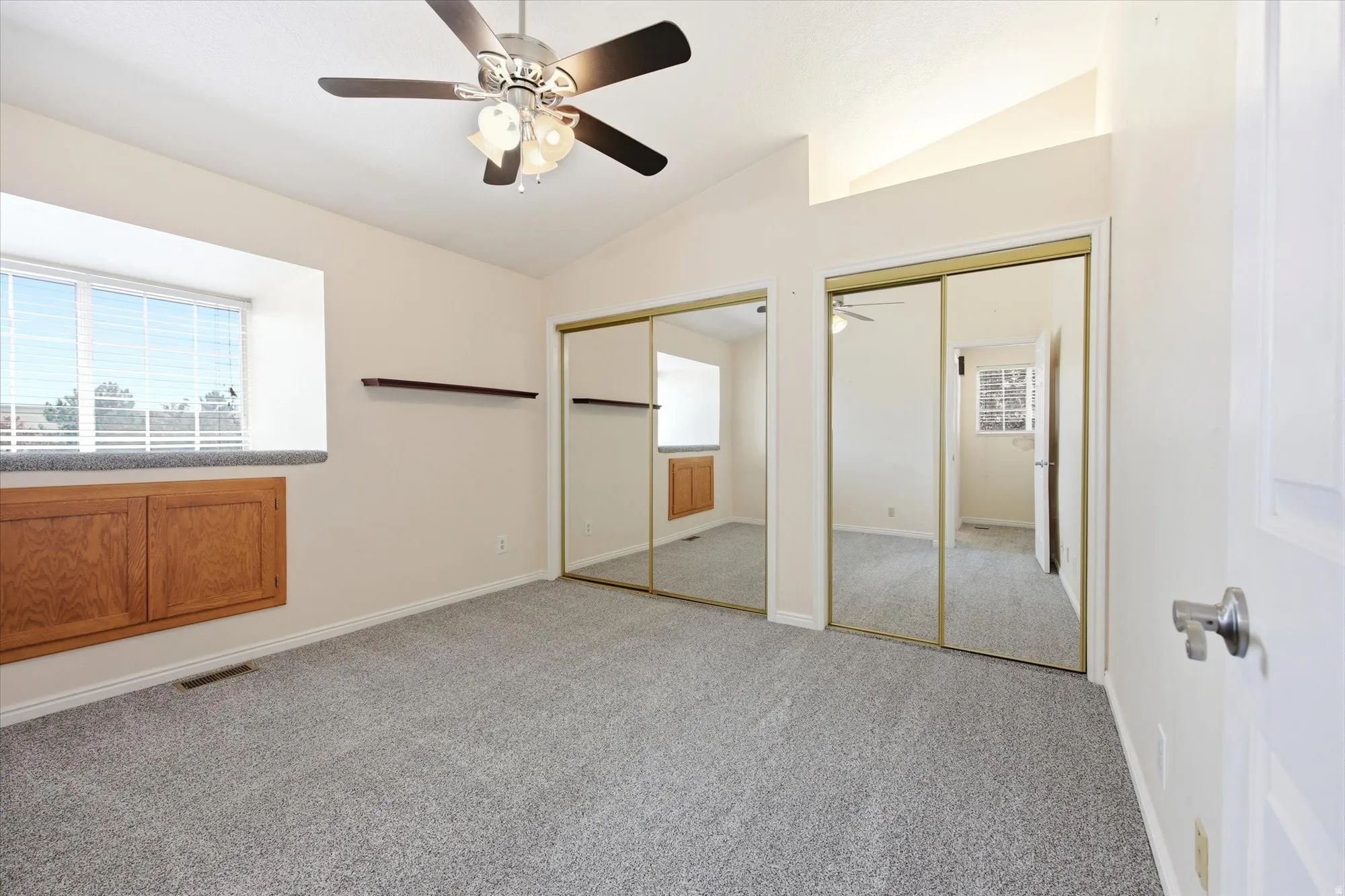 Spacious room featuring vaulted ceilings, a ceiling fan with integrated lighting, built-in lower cabinetry with wood-finish doors, mirrored closet doors, and light gray carpeting