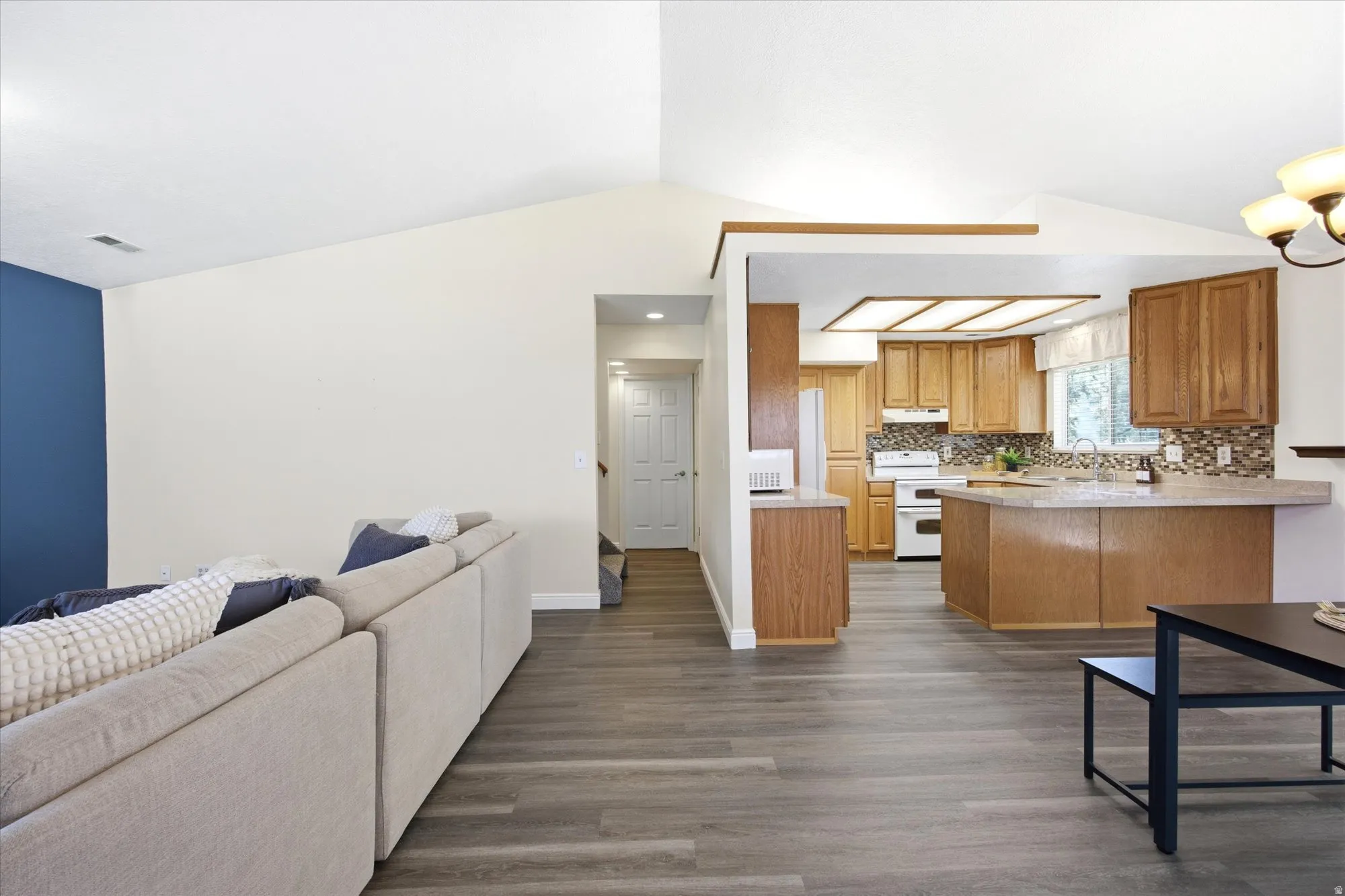 Open-concept living area featuring wood-finish flooring, vaulted ceilings, and a kitchen with wood cabinetry, a tile backsplash, and a breakfast bar