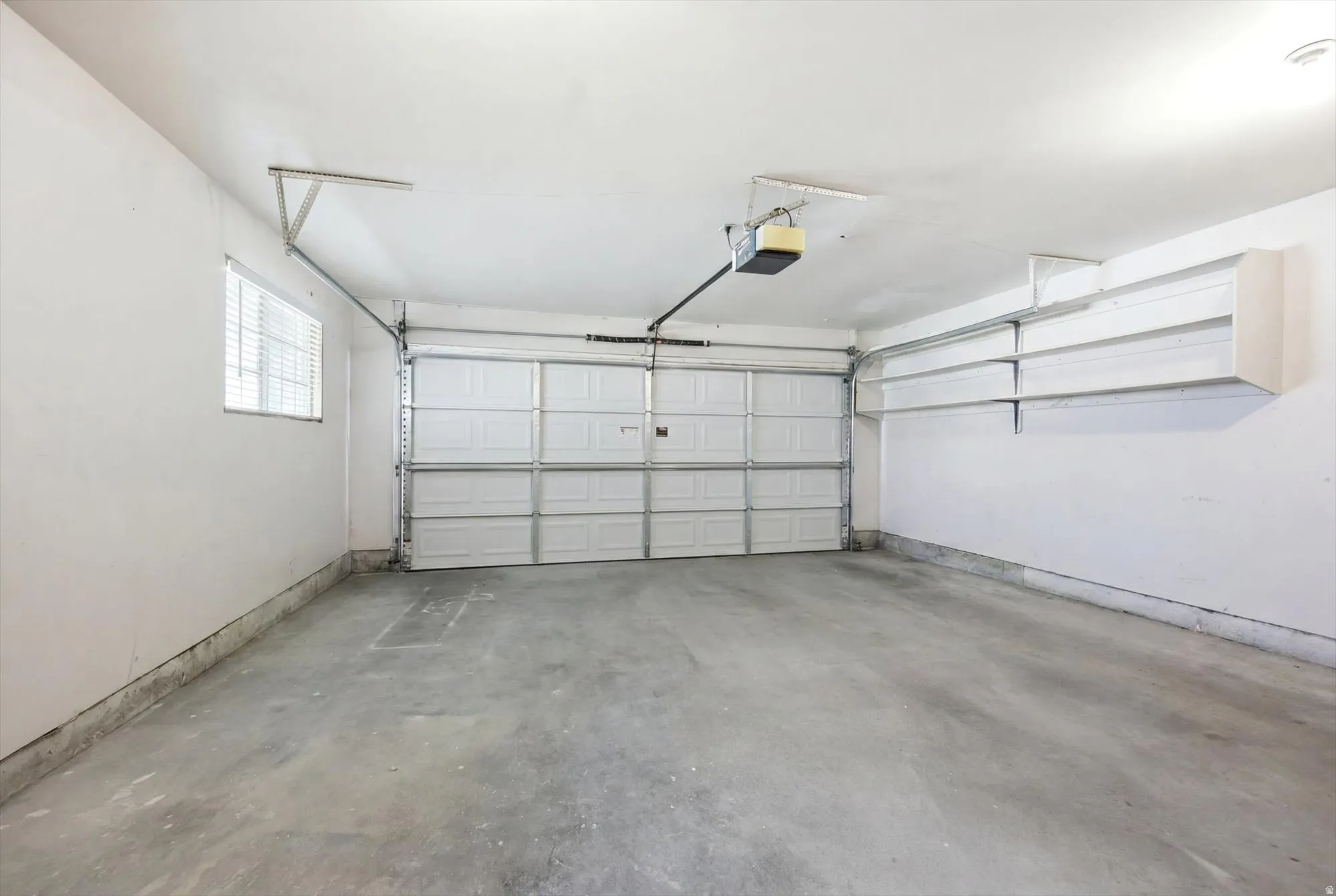 Attached two-car garage featuring a paneled garage door, a side window with horizontal blinds, built-in shelving, and a concrete floor