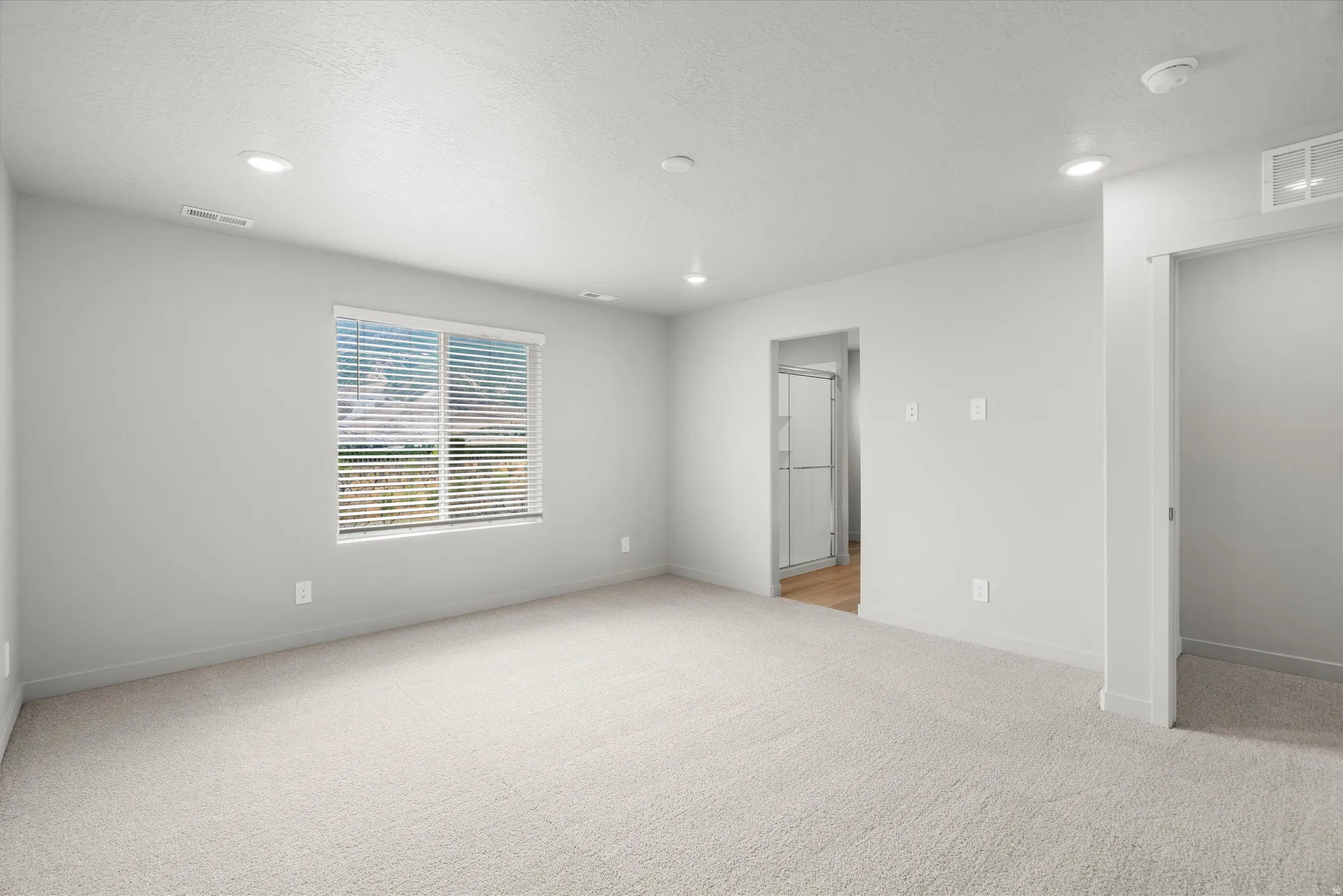 Bright room featuring light-toned carpeting, recessed ceiling lighting, a window with horizontal blinds, and a doorway revealing wood-finish flooring