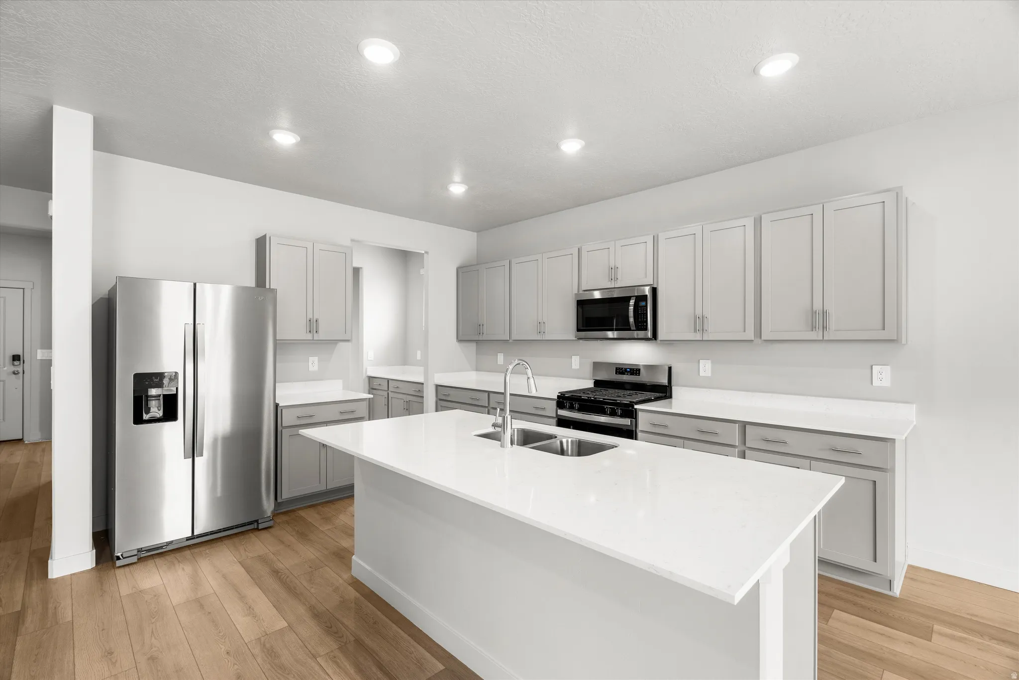 Open-concept kitchen featuring an island with an undermount sink, stainless steel appliances, light gray shaker cabinetry, white countertops, and wood-finish flooring