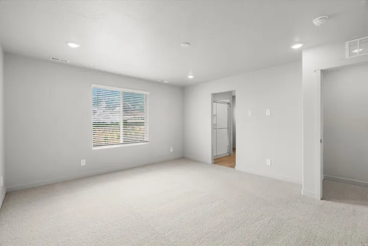 Bright room featuring light-toned carpeting, recessed ceiling lighting, a window with horizontal blinds, and a doorway revealing wood-finish flooring