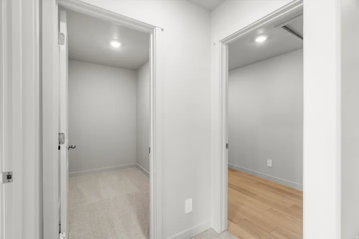 Hallway with two open doorways, one revealing a carpeted room with an overhead light, and the other showing a room with wood-finish flooring and a recessed light