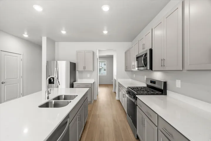 Gourmet kitchen featuring an expansive island with double stainless steel sink, light wood-finish flooring, recessed lighting, sleek gray cabinetry, and stainless steel appliances