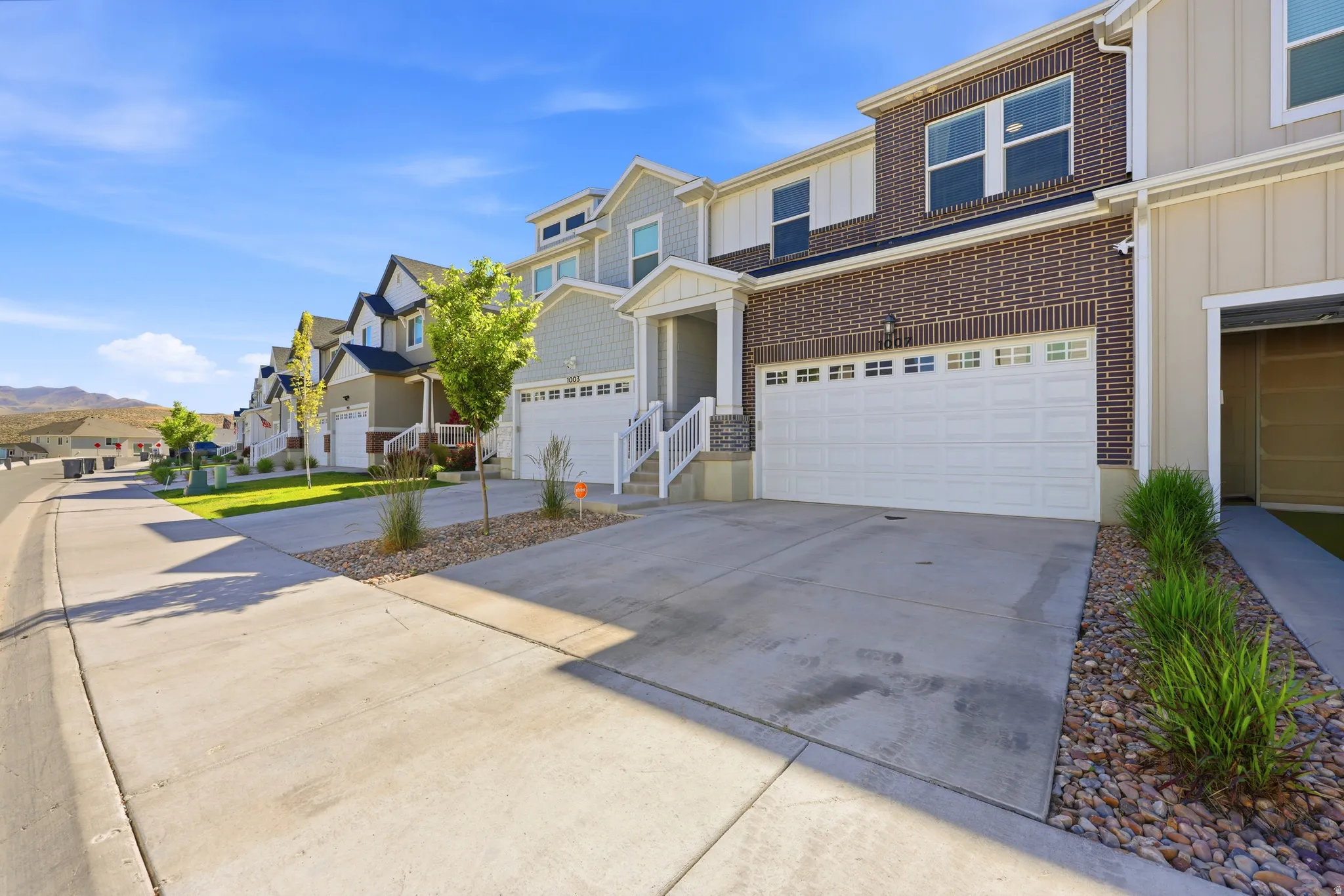 Contemporary townhome exterior featuring brick and board-and-batten siding, a two-car garage, concrete driveway, and low-maintenance landscaping with decorative rock and mature plantings