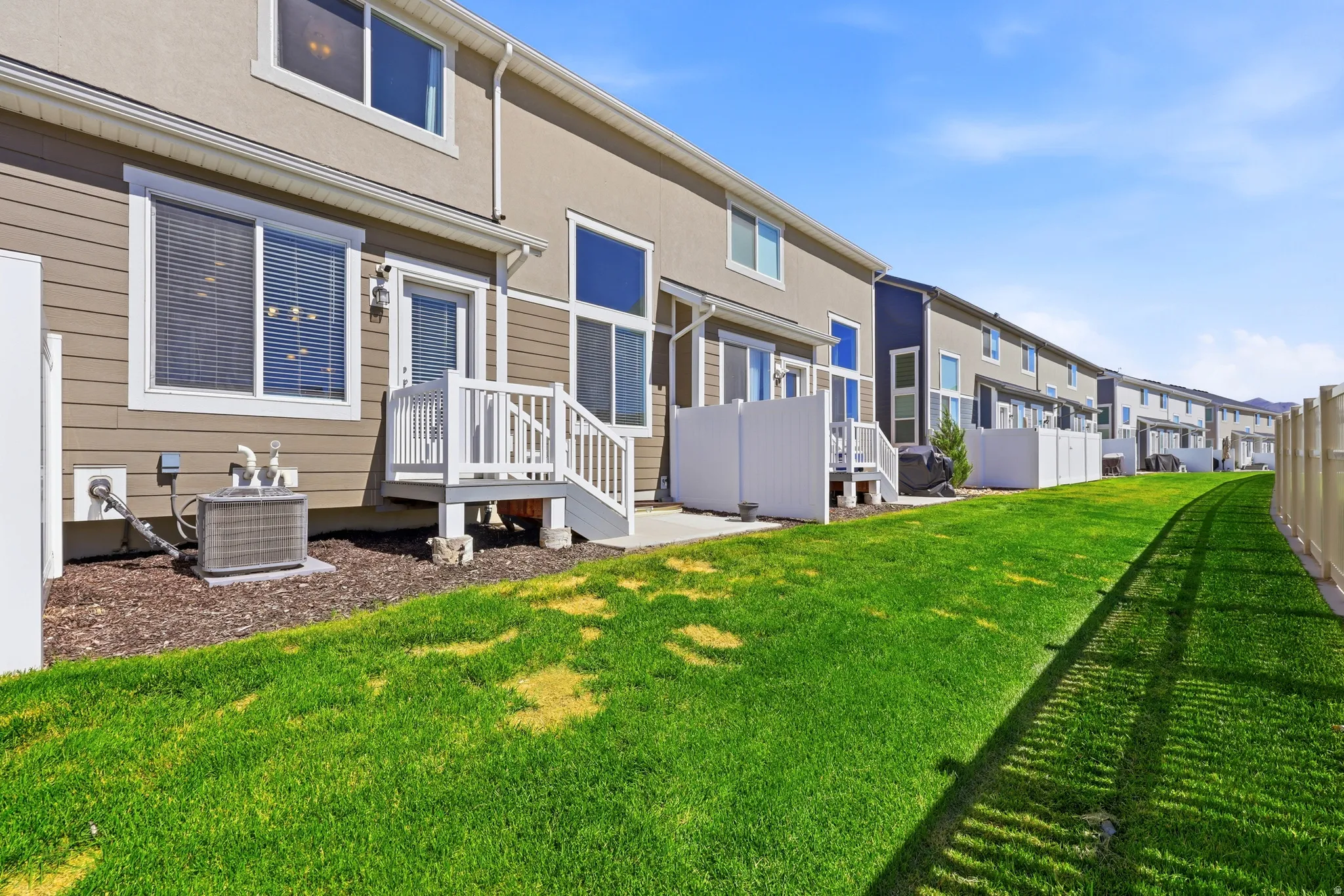 Expansive green space featuring a manicured lawn, individual back patios with white railings, tan and brown siding, and white vinyl fencing