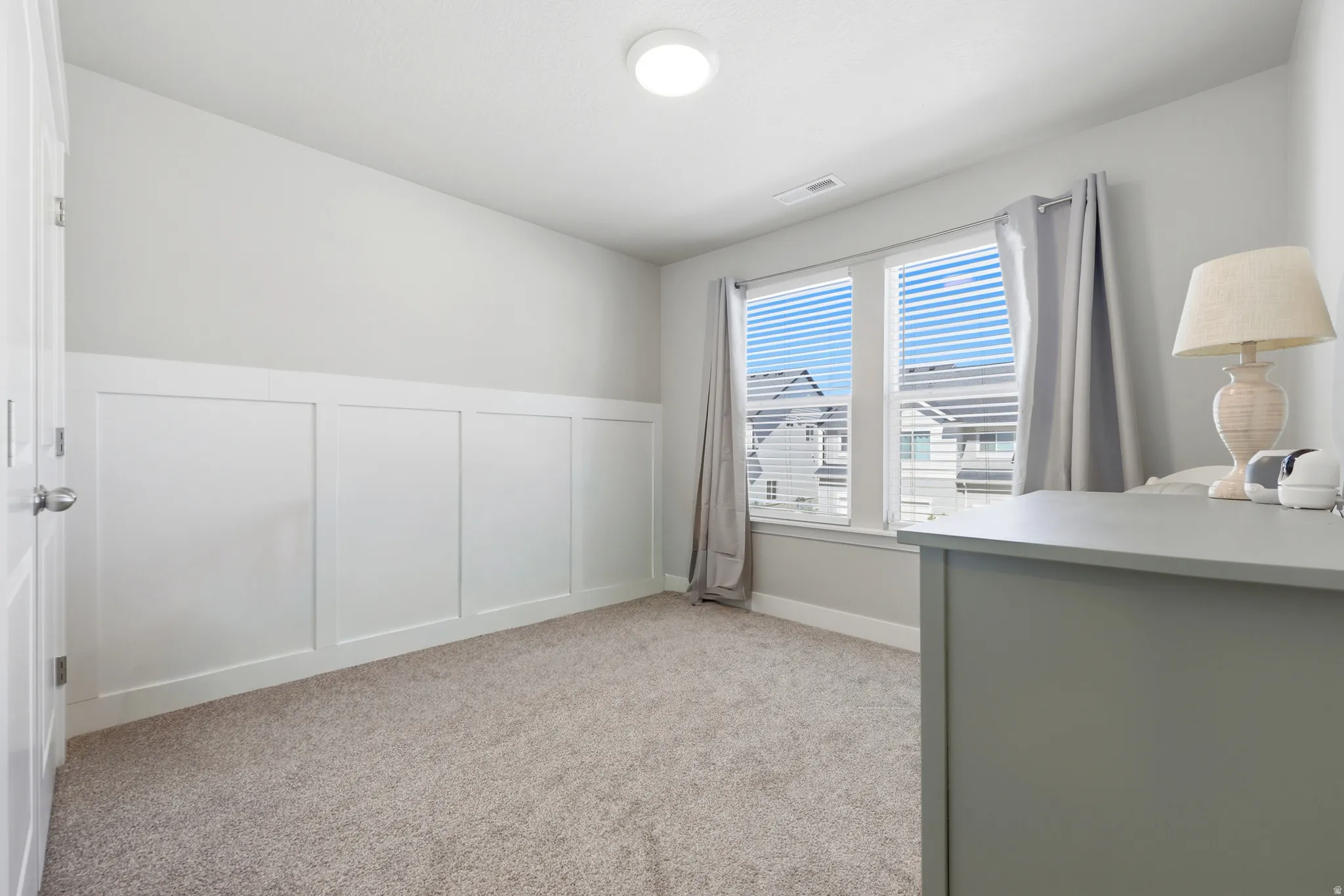 Room featuring carpet flooring, two windows with blinds and curtains, and wainscoting wall detailing