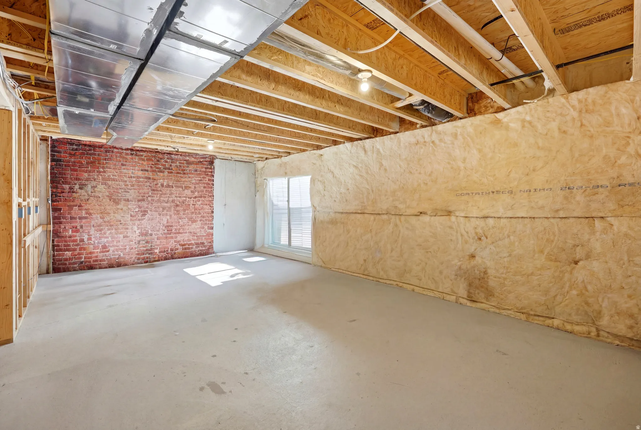 Unfinished basement space featuring exposed brick, concrete flooring, exposed ceiling joists, and a window