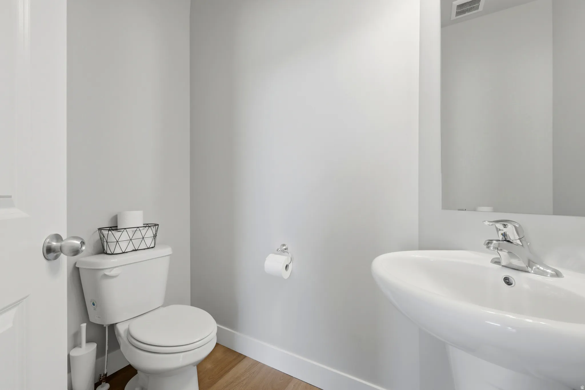 Pedestal sink with chrome faucet, toilet with wood-finish flooring, and light gray walls