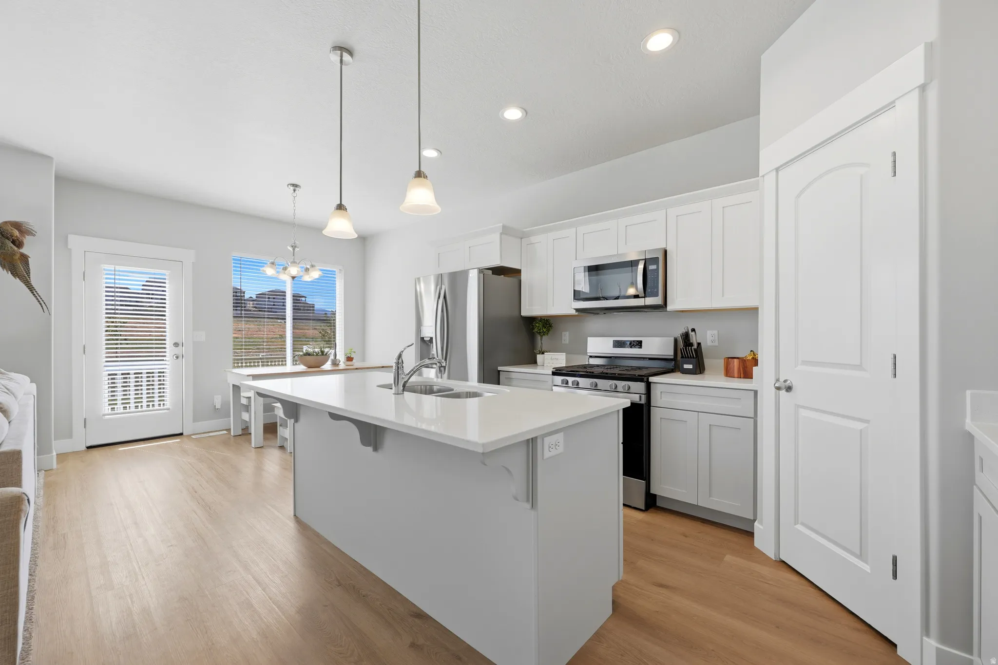 Modern kitchen featuring an expansive island with a built-in sink and pendant lighting, white cabinetry with stainless steel appliances, wood-finish flooring, a dining area with a chandelier, and a glass-paneled exterior door