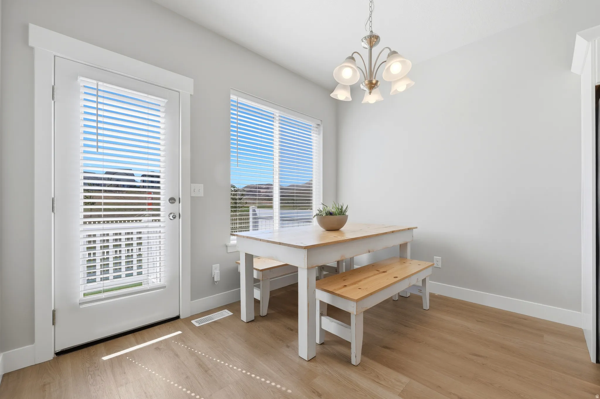 Light-filled dining area featuring wood-finish flooring, a contemporary chandelier, a large window with blinds, a glass-paned door with blinds, and white baseboards