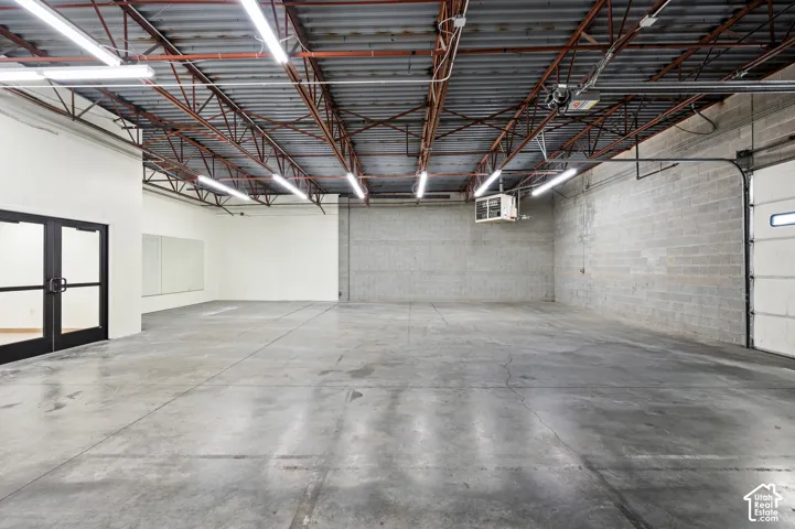 Spacious commercial interior featuring concrete flooring, exposed ceiling trusses, fluorescent overhead lighting, cinder block walls, and a large roll-up door