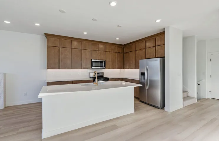 Modern kitchen featuring a white island with a gold-finish faucet, stainless steel appliances, wood-finish cabinetry, and white countertops with under-cabinet lighting