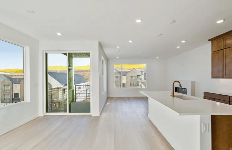 Open-concept living area featuring light wood-finish flooring, a white kitchen island with an integrated sink and gold-tone faucet, recessed ceiling lighting, and a sliding glass door opening to a private balcony with metal railings