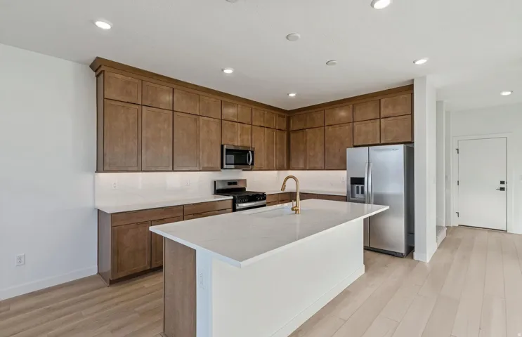 Open-concept kitchen featuring an expansive island with white countertop, rich wood-finish cabinetry, stainless steel appliances, and light wood-finish flooring