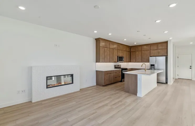 Open-concept living space featuring a modern linear fireplace with a herringbone tile surround, wood-finish flooring, recessed lighting, and a kitchen with rich wood cabinetry, stainless steel appliances, and a central island with a sink and white counter