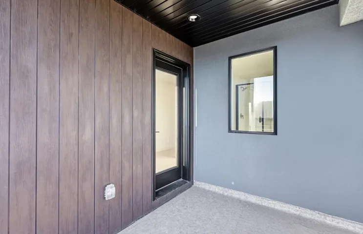 Exterior patio featuring wood-finish paneling, a full-lite glass door with dark trim, a fixed window with dark trim, a dark soffit with recessed lighting, and light gray stucco