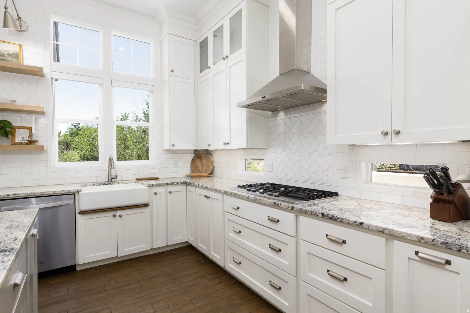 Bright kitchen featuring white shaker cabinetry, patterned stone countertops, stainless steel appliances, a white farmhouse sink, and wood-finish flooring