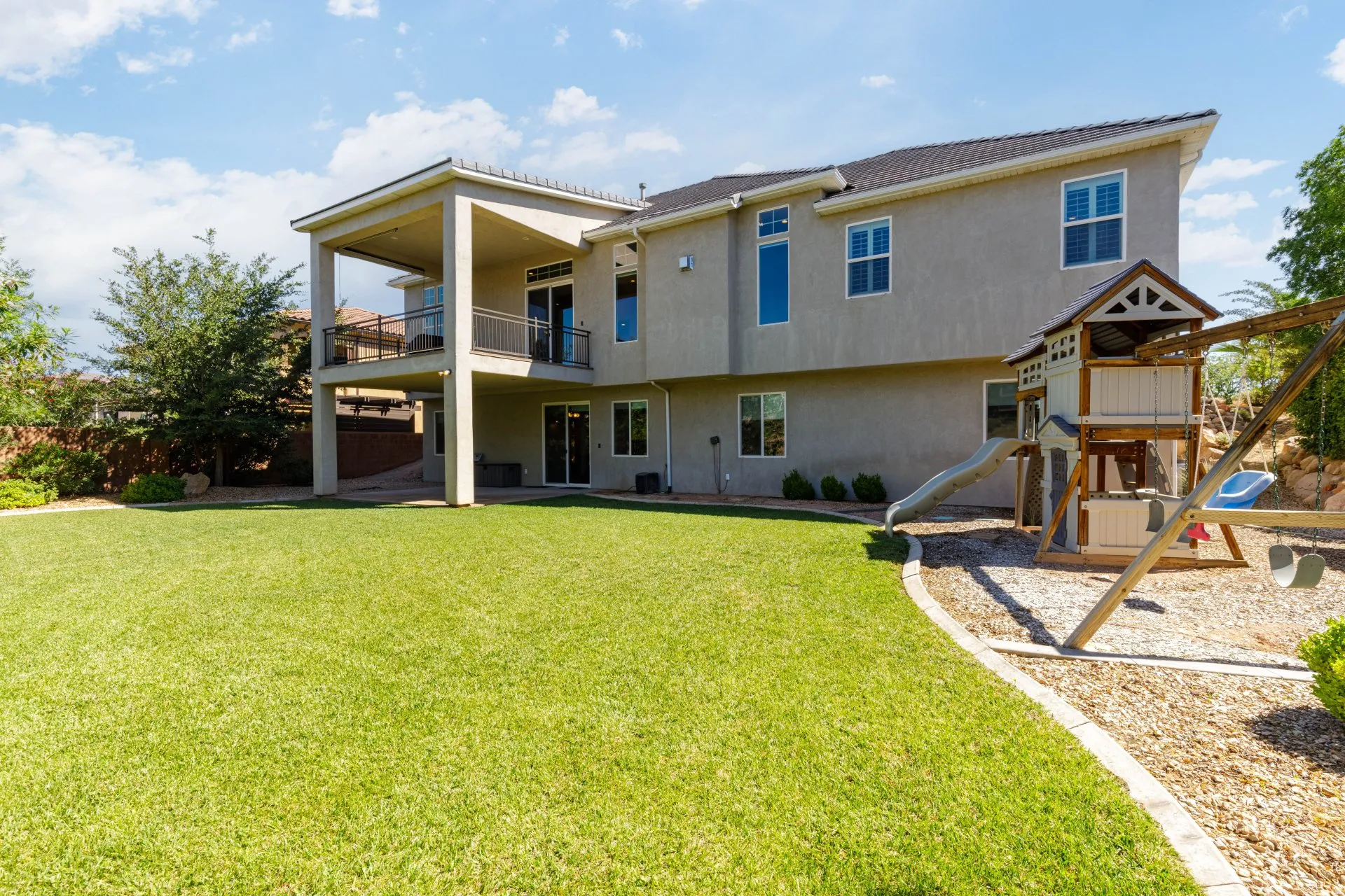 Expansive private backyard featuring a covered patio, upper-level balcony with metal railings, a manicured lawn, and a designated play area with wood chip ground cover