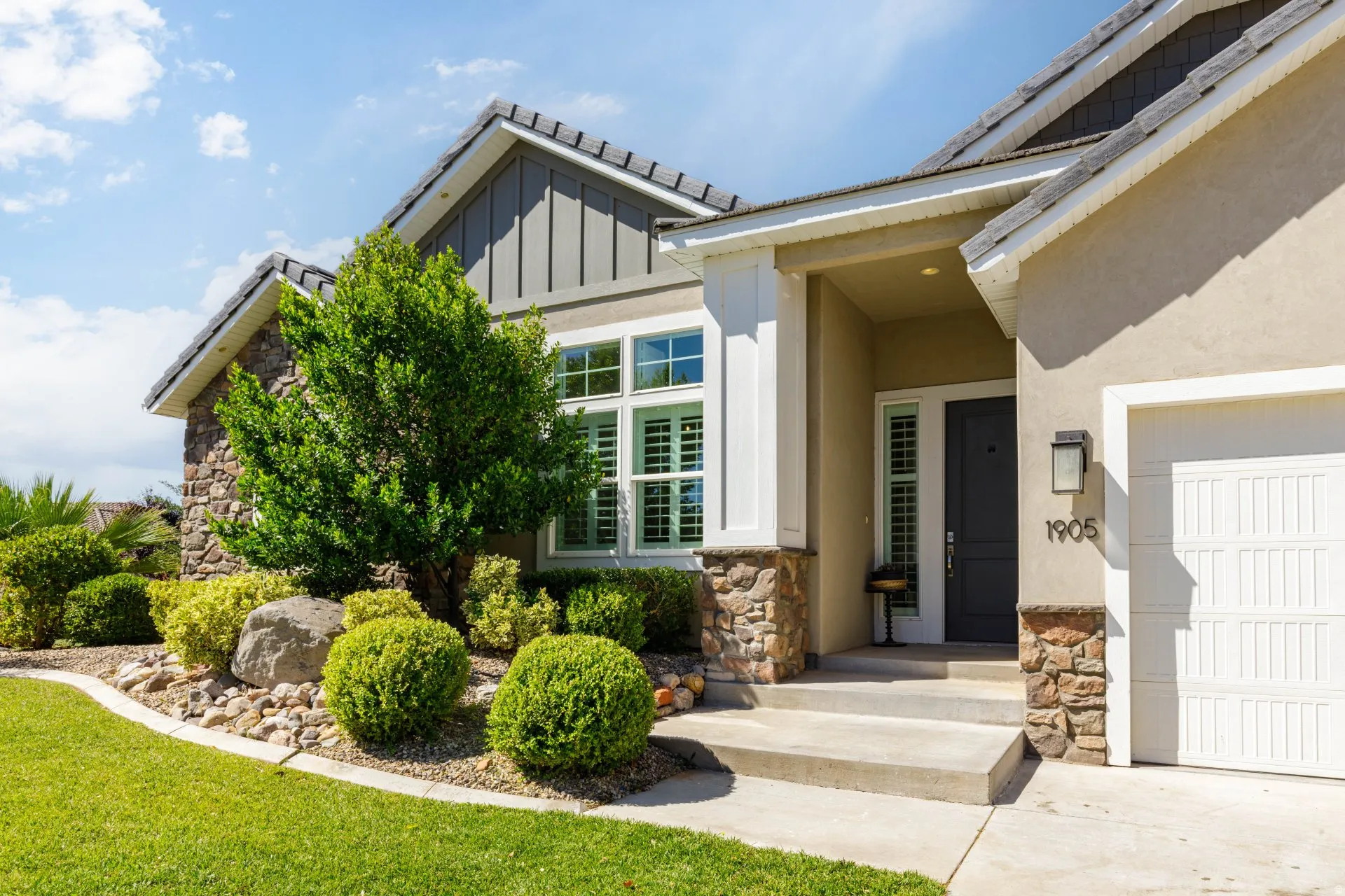 Stucco and stone exterior featuring a dark gray front door, a white garage door, and a landscaped front yard with green shrubs and a mature tree