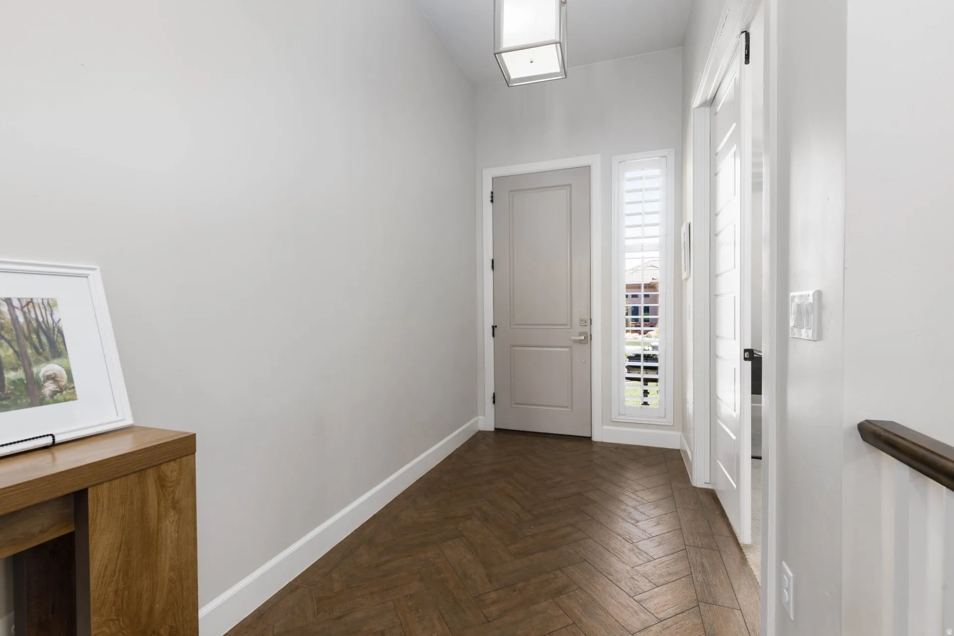Entryway featuring wood-finish herringbone flooring, a neutral-toned door, a vertical window with white plantation shutters, a square ceiling-mounted light fixture, and white baseboards