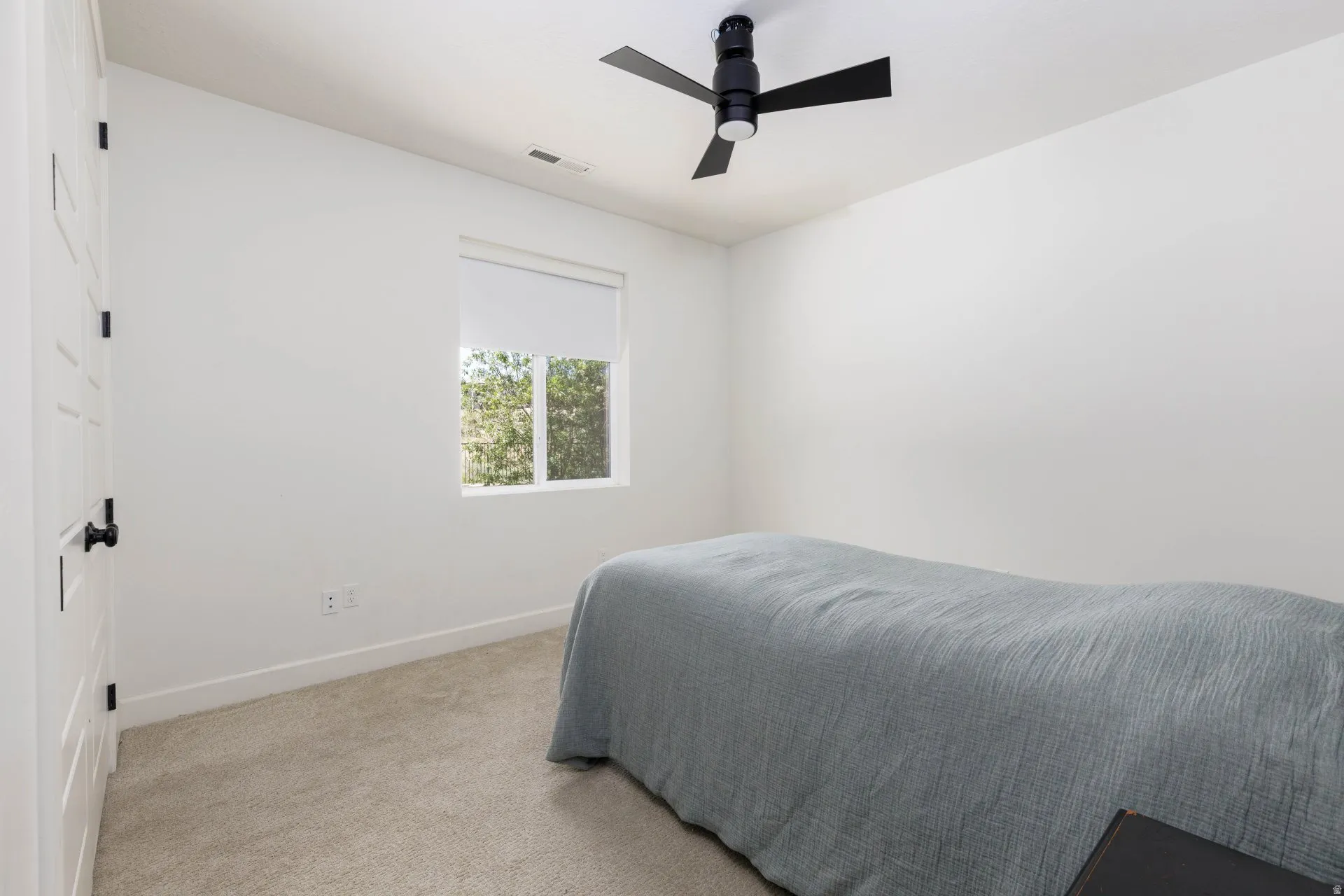 Bright room with a neutral color palette featuring a window with a roller shade, light carpet flooring, and a modern black ceiling fan