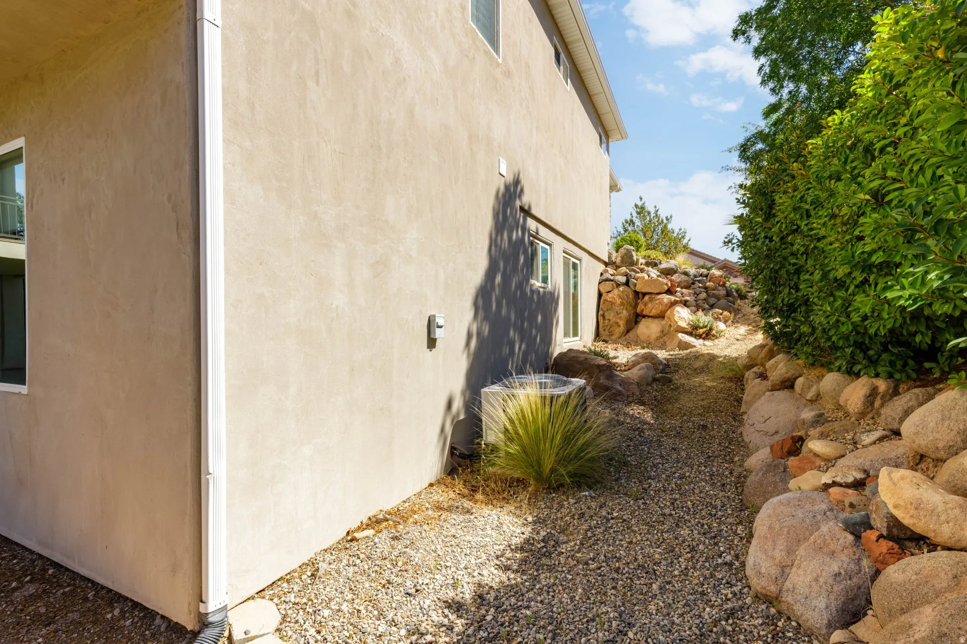 Exterior stucco finish, with gravel landscaping, a rock retaining wall, established shrubbery, and an outdoor HVAC unit