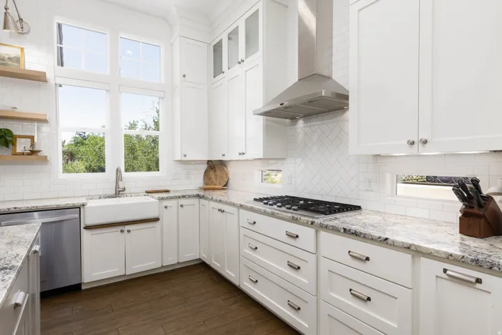 Bright kitchen featuring white shaker cabinetry, patterned stone countertops, stainless steel appliances, a white farmhouse sink, and wood-finish flooring