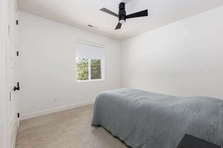 Bright room with a neutral color palette featuring a window with a roller shade, light carpet flooring, and a modern black ceiling fan