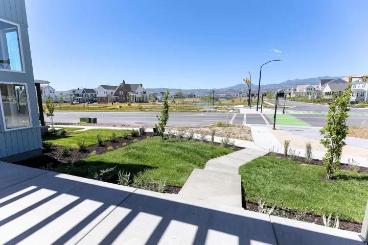 Front porch with concrete flooring and slatted railing shadows, surrounded by manicured lawn and landscaping, with a sidewalk leading to a paved street featuring traffic signals and painted crosswalks, set against a backdrop of distant mountains