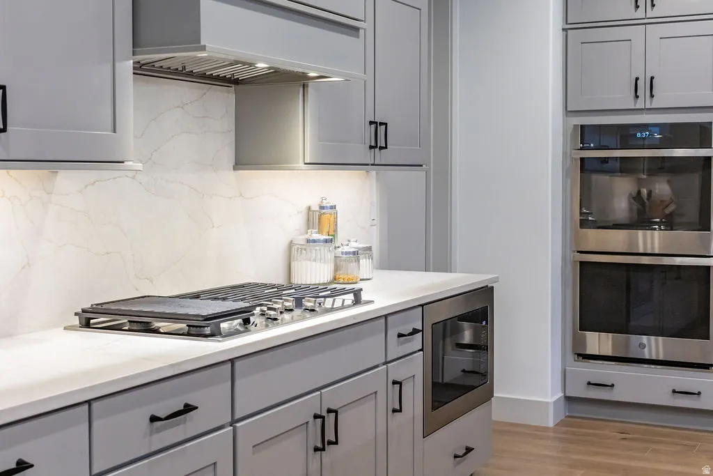 Modern kitchen featuring light gray cabinetry with matte black hardware, white countertops, a built-in gas cooktop, a stainless steel range hood, and a built-in double oven