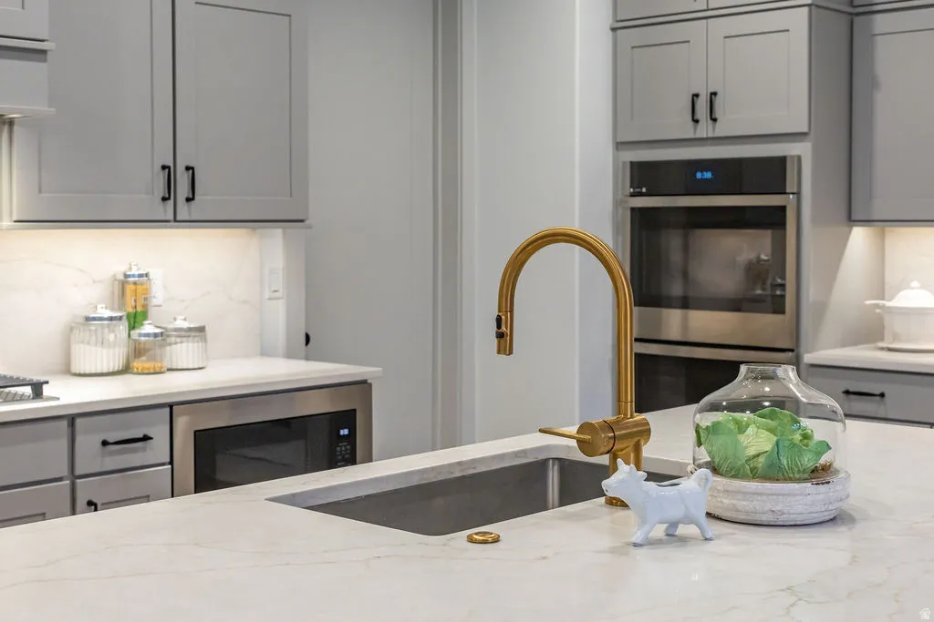 Kitchen featuring light gray cabinetry with dark hardware, a brass-finish gooseneck faucet, a double wall oven, a built-in microwave, and light-toned countertops with an integrated sink