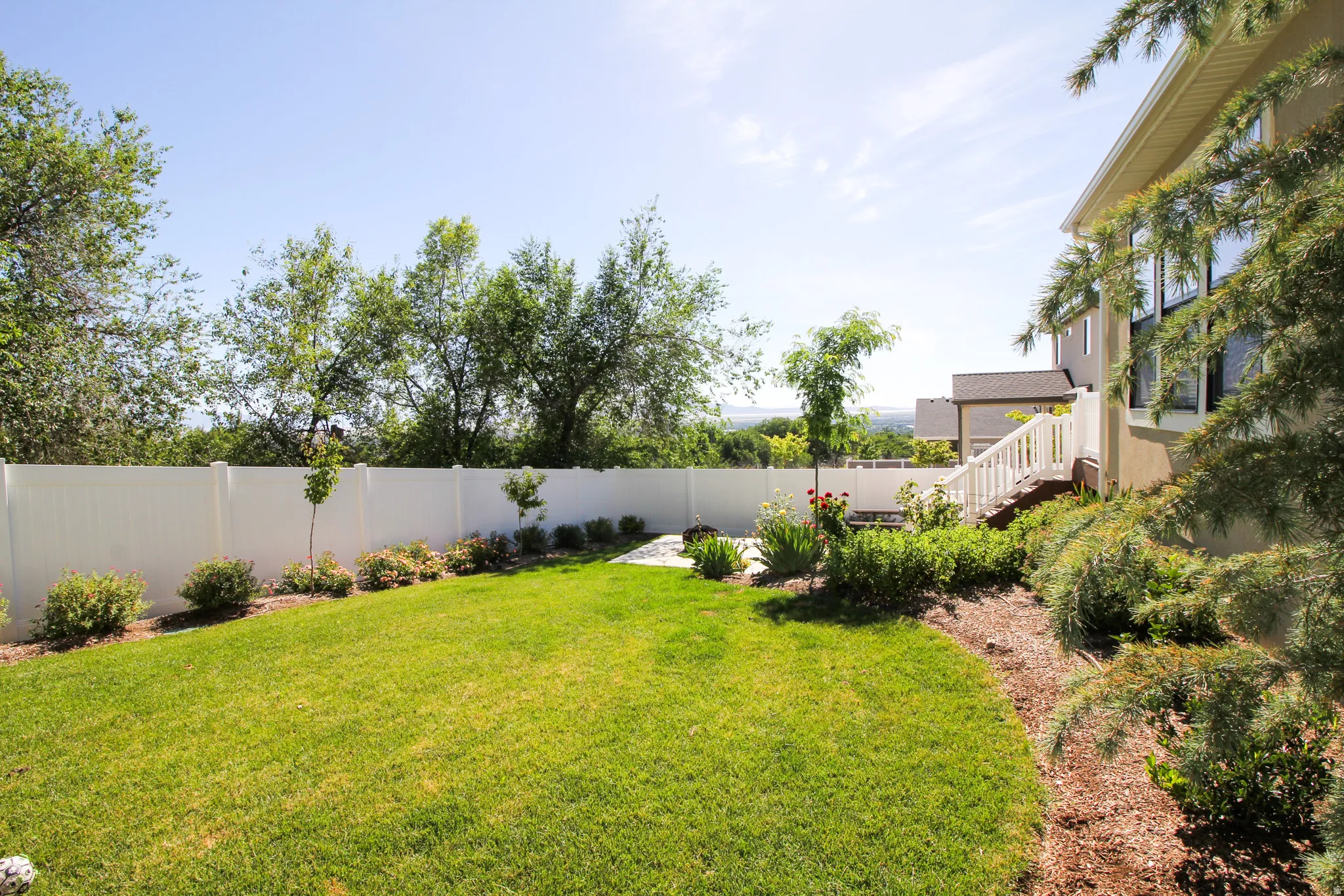 Expansive backyard featuring a manicured lawn, a white privacy fence, mature trees, a multi-level deck with white railings, and integrated landscaping