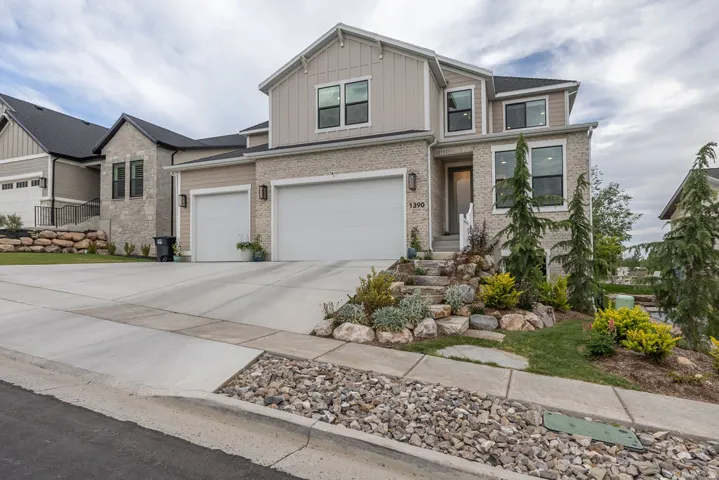Modern residential exterior featuring stone and board-and-batten siding, a two-car garage, a concrete driveway, and a landscaped rock garden with established plantings