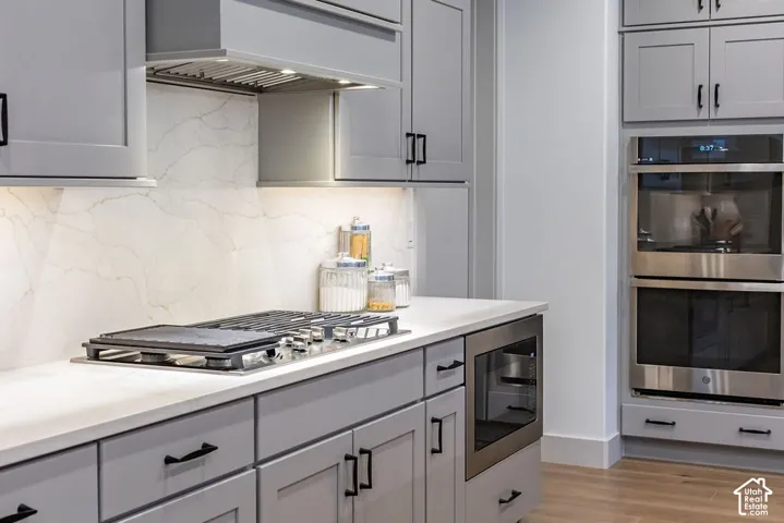 Modern kitchen featuring light gray cabinetry with matte black hardware, white countertops, a built-in gas cooktop, a stainless steel range hood, and a built-in double oven