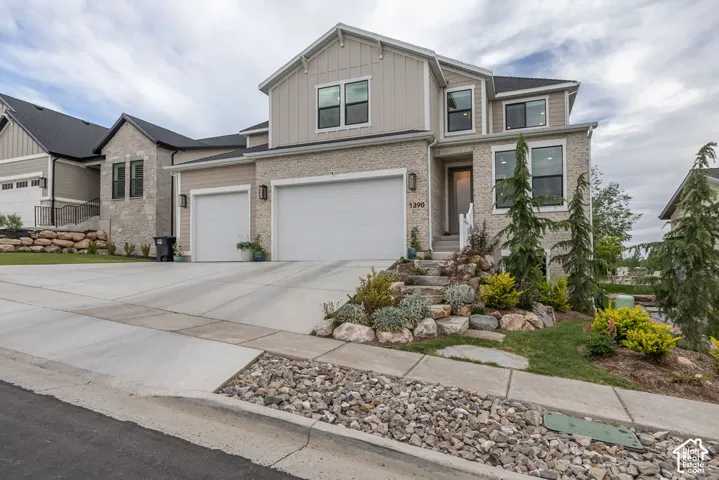 Modern residential exterior featuring stone and board-and-batten siding, a two-car garage, a concrete driveway, and a landscaped rock garden with established plantings