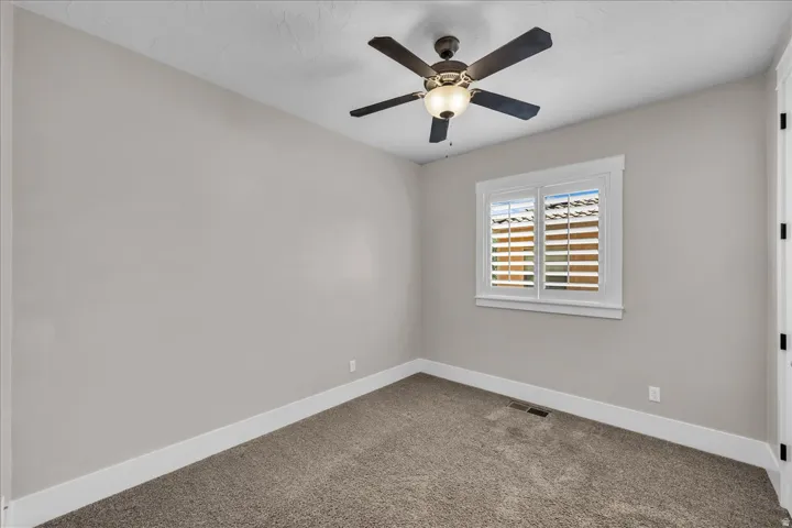Neutral-toned room featuring plush carpet flooring, a window with plantation shutters and white trim, a dark-bladed ceiling fan with integrated lighting, and white baseboards