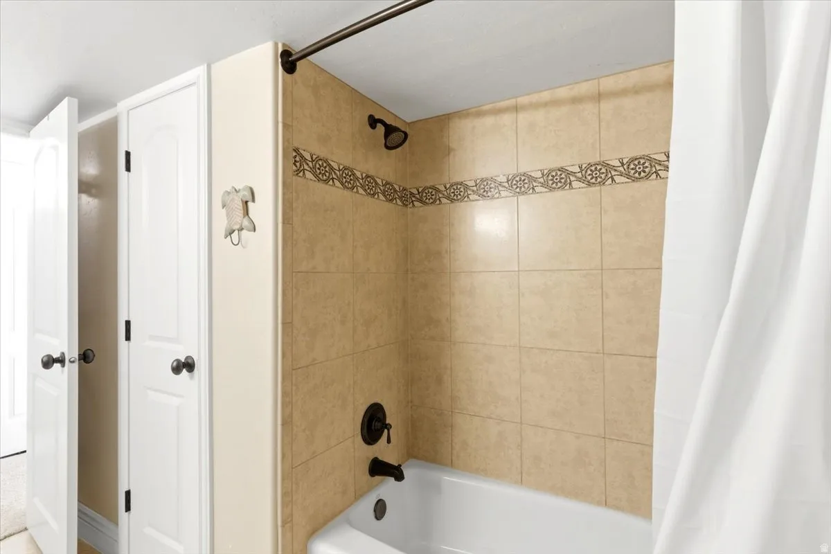 Bathtub and shower combination featuring beige tile surround with decorative accent band, dark bronze fixtures, and an adjacent linen closet with bi-fold doors