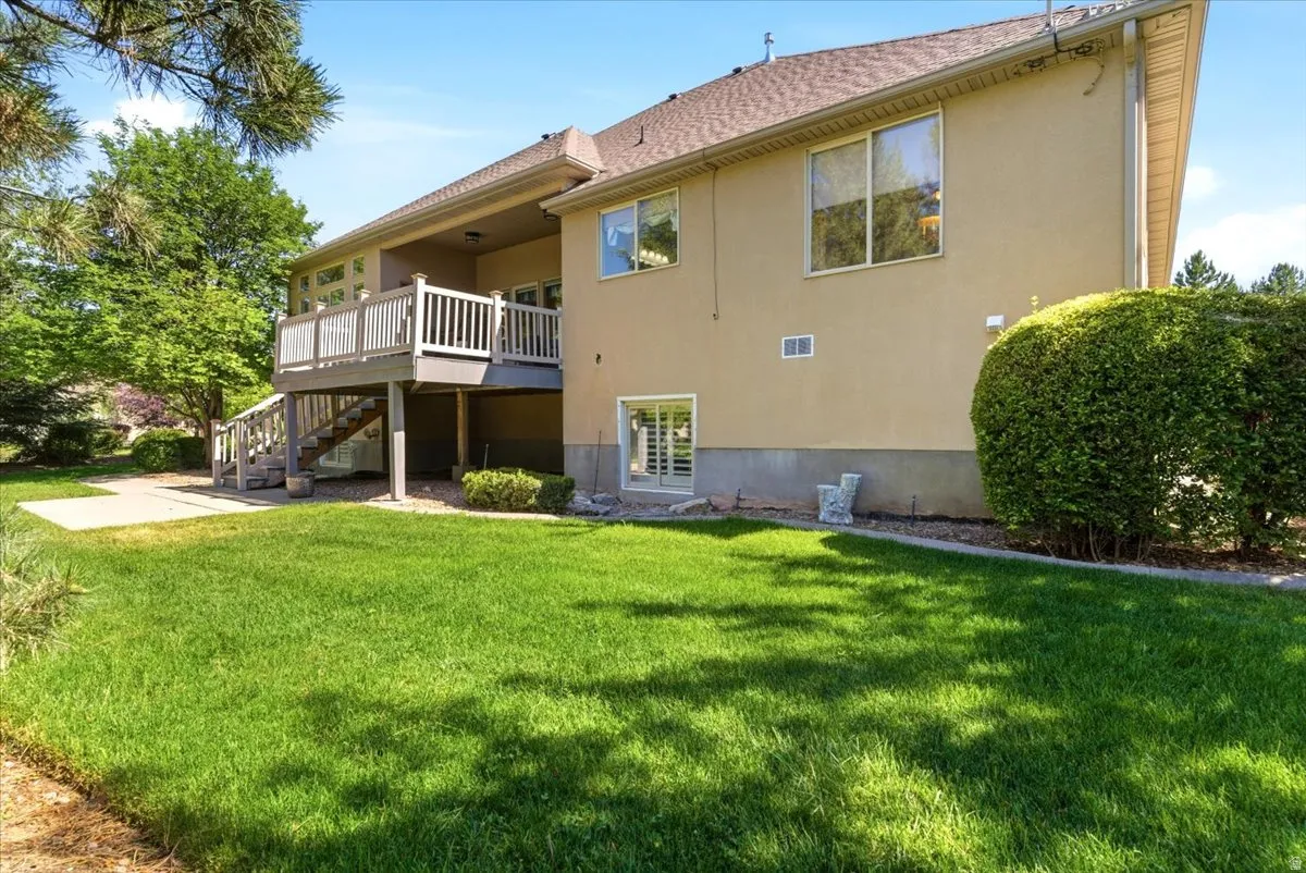 Expansive backyard featuring a manicured lawn, a raised wood-finish deck with white railings, exterior stairs, a lower-level concrete patio, and a sliding glass door entry