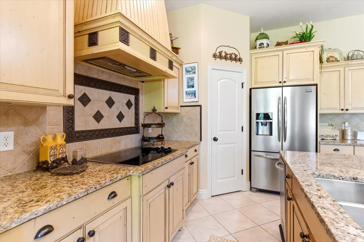 Kitchen featuring light-toned cabinetry with dark hardware, granite countertops, a tiled backsplash with decorative accents, an electric cooktop, and a stainless steel refrigerator