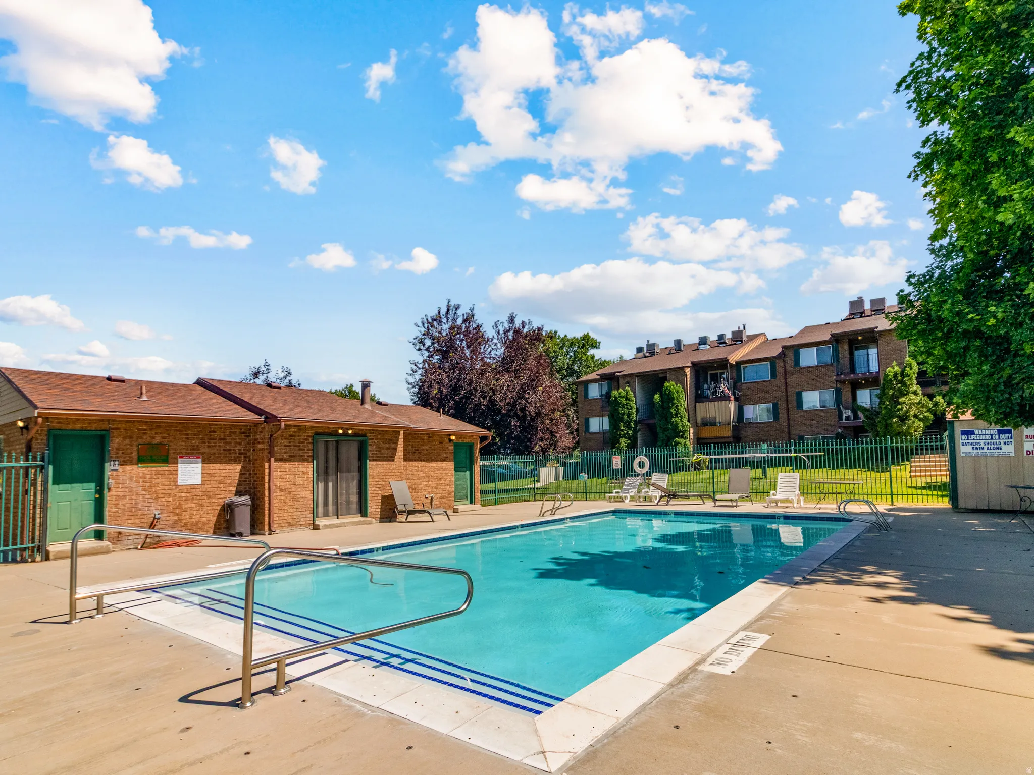 Outdoor swimming pool with concrete surround, metal handrails, and a brick pool house featuring green doors